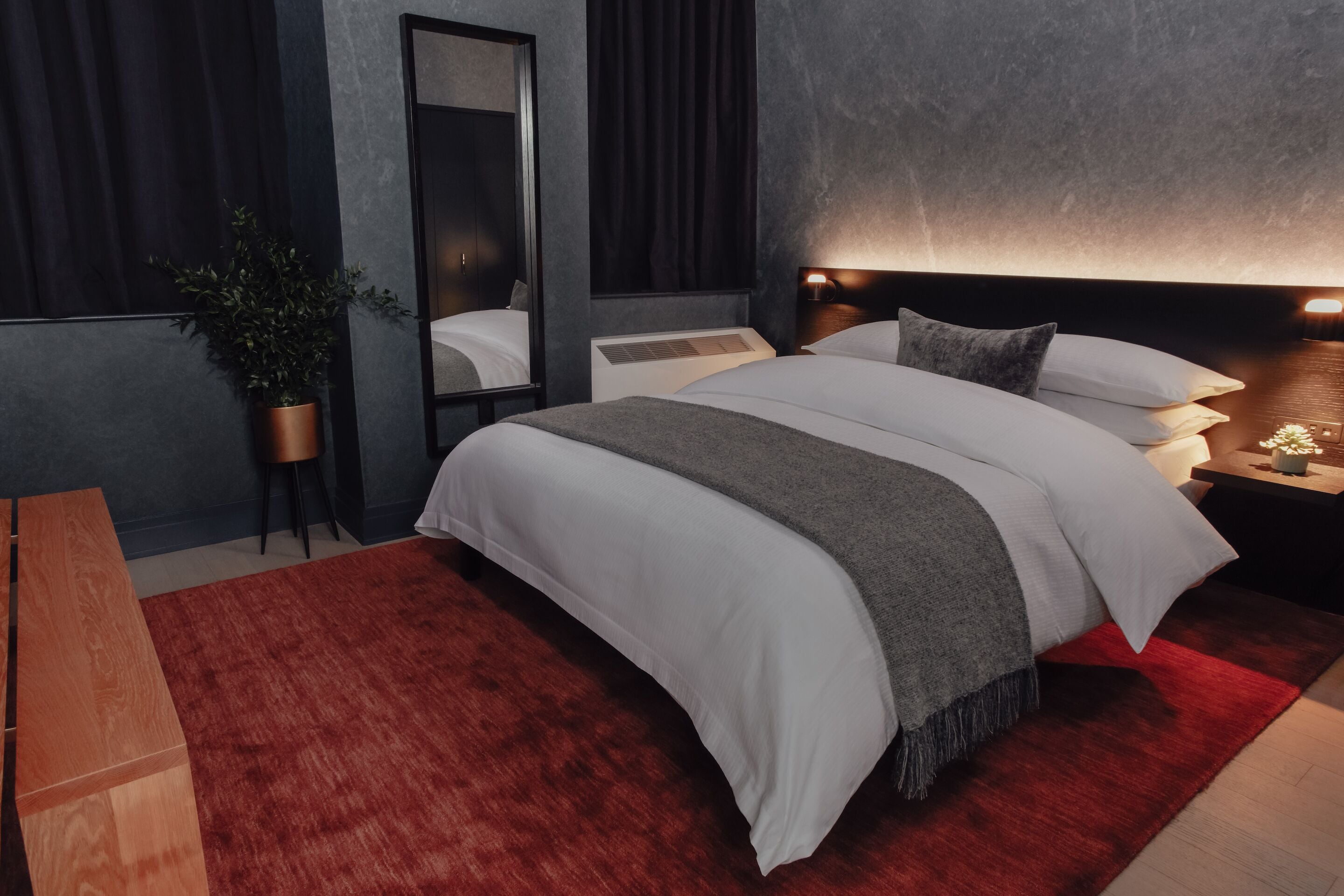 A modern bedroom with a large bed, a bedside table, a nightstand, a mirror, and a red rug.