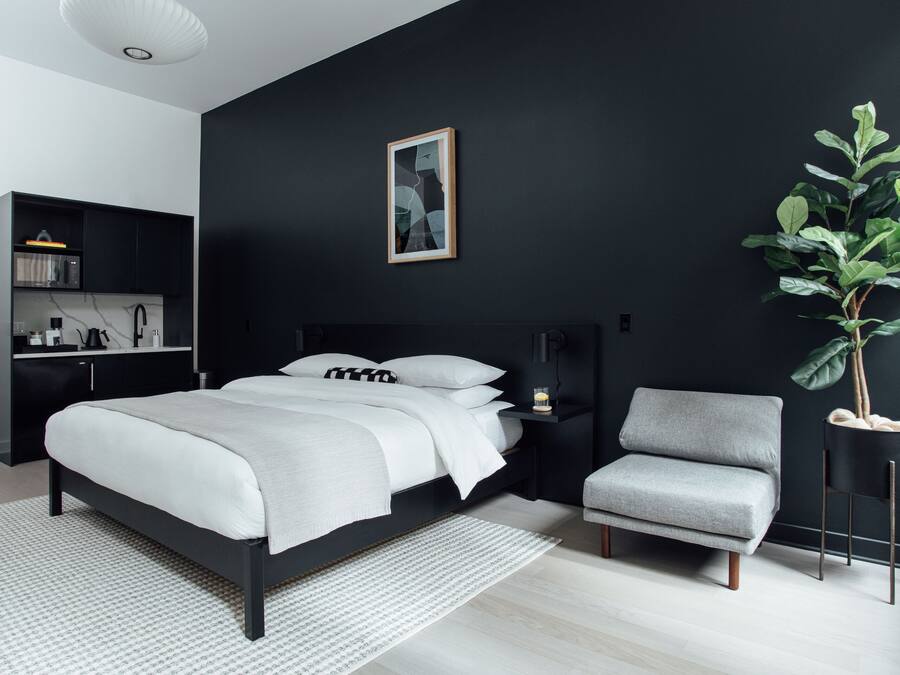 Una camera da letto moderna con una parete nera, un letto con biancheria da letto bianca, una poltrona grigia, un comodino con una lampada e una pianta.