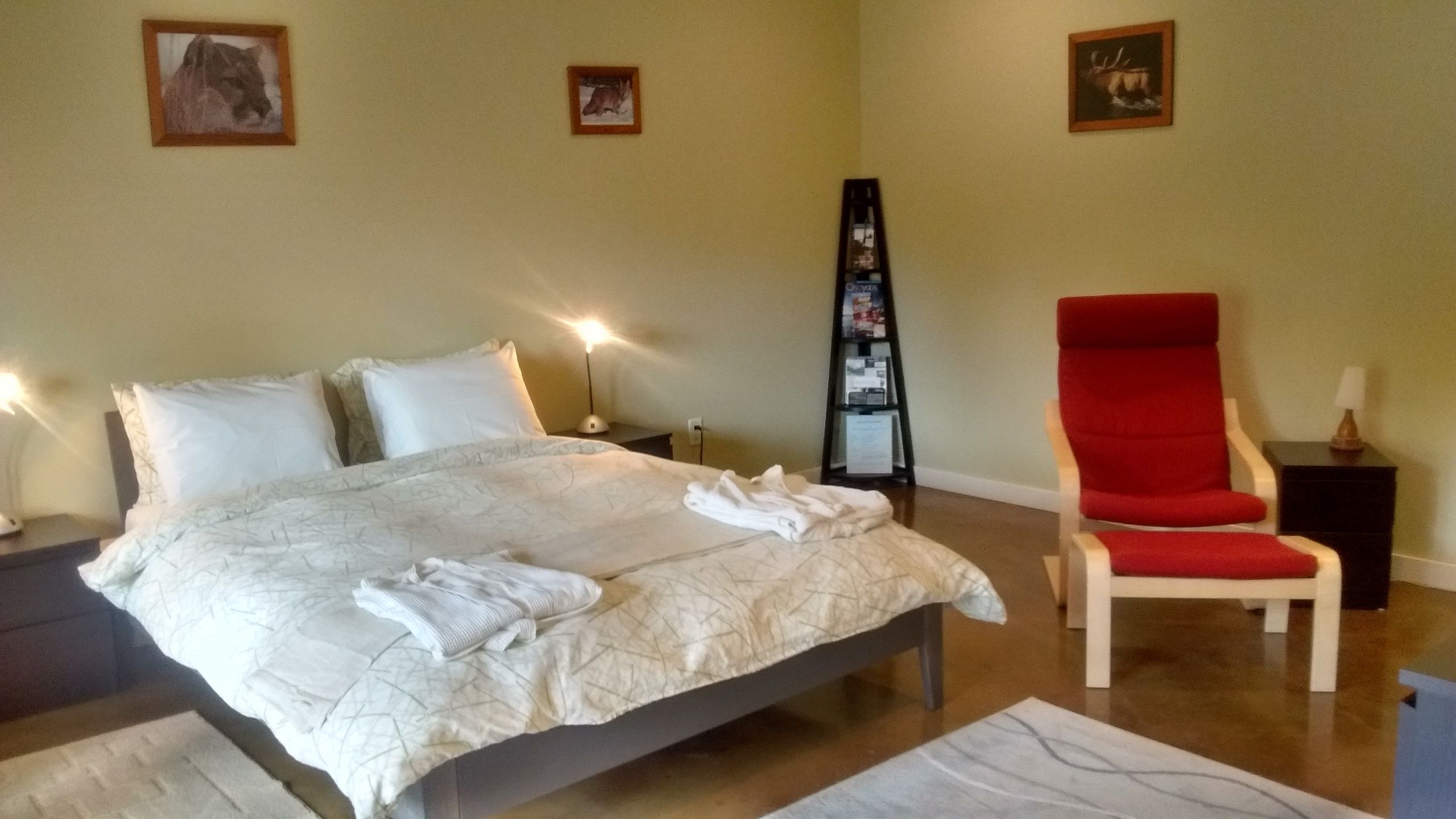 Een opgemaakt bed met witte lakens en handdoeken, een rode fauteuil en een klein tafeltje met een lamp.