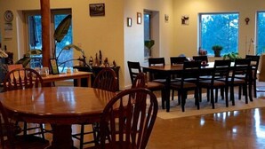 A dining area with wooden tables and chairs, a large window with a view of trees, and a plant on the windowsill. - Baergnaescht Bed & Breakfast (Osoyoos)