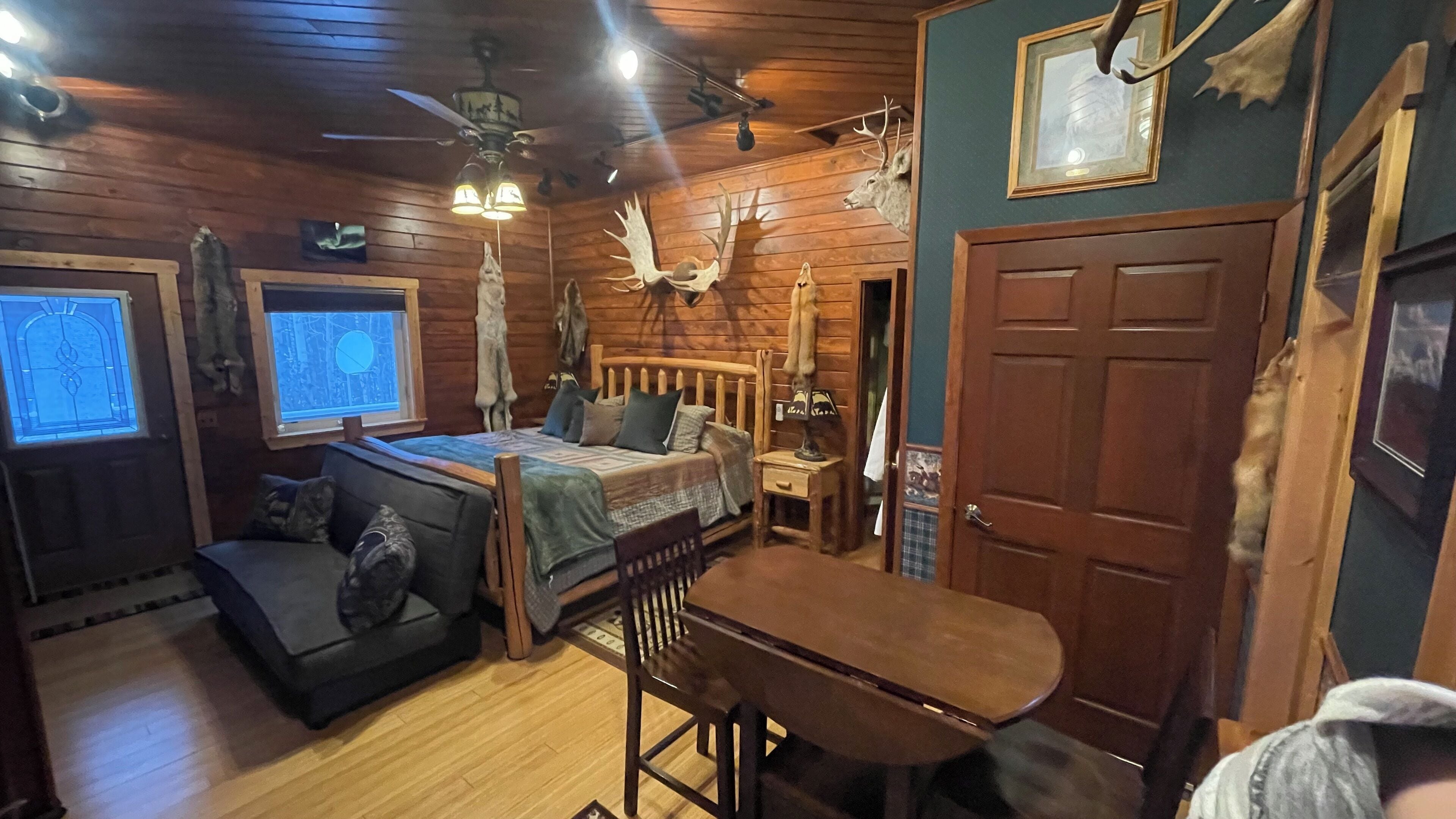 A cozy cabin-style room with a bed, a sofa, a dining table, and a ceiling fan.