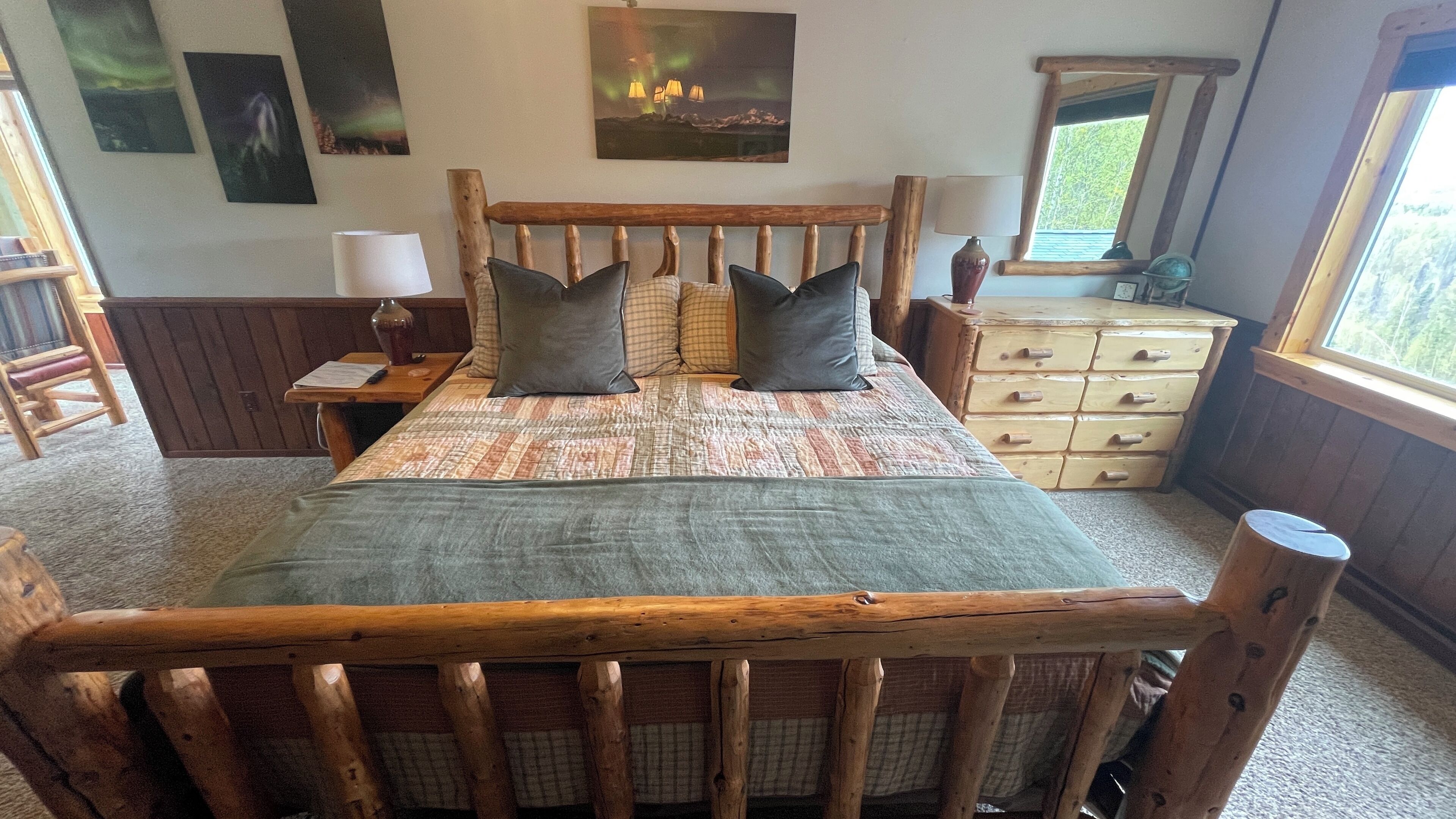 A wooden-framed bed with a quilt and pillows, a nightstand with a lamp, a dresser with drawers, and a window with a view of trees.