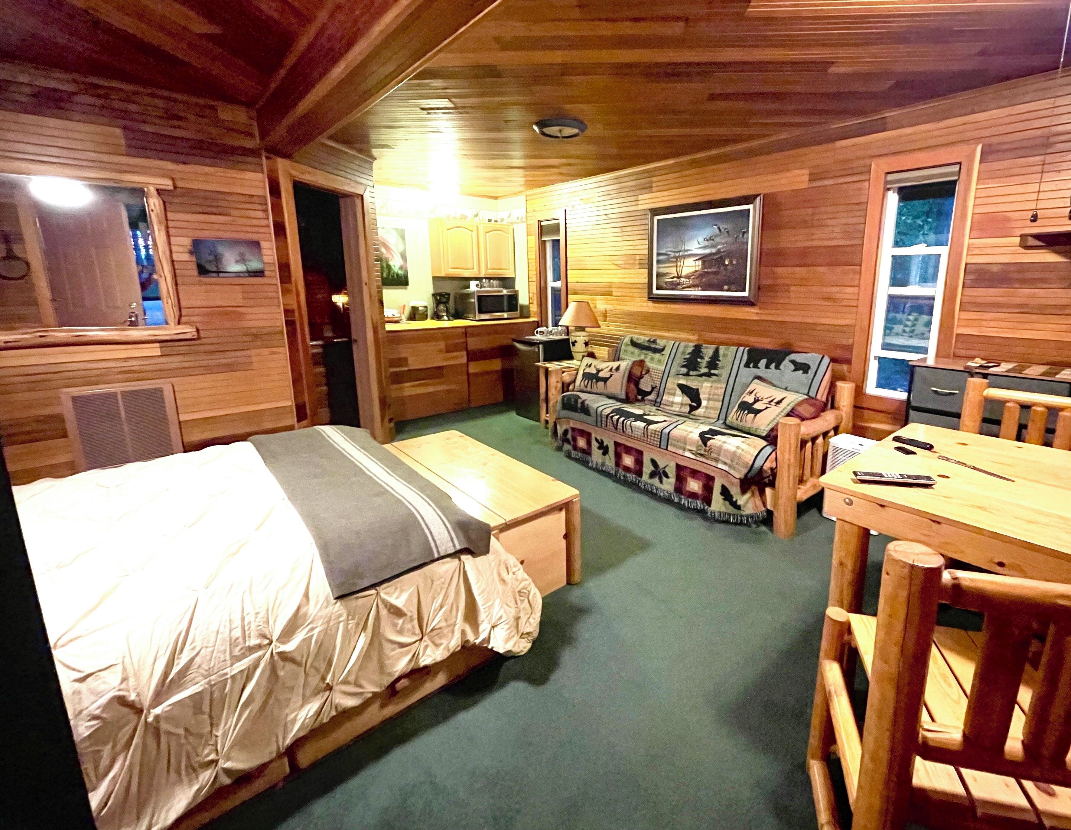 A cozy cabin-style room with a bed, a sofa, a dining table, and a kitchenette.