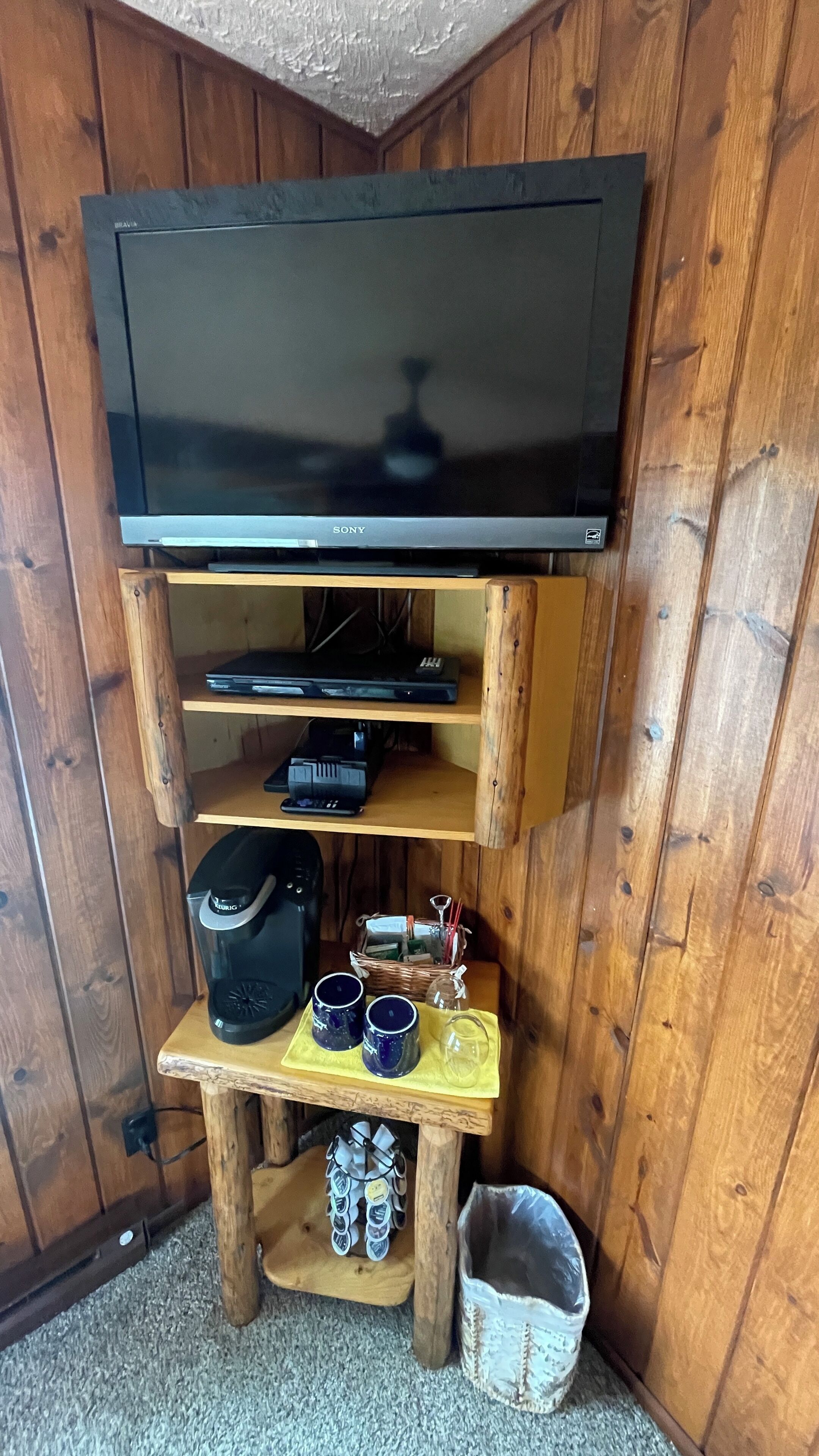 A wooden entertainment unit with a flat-screen TV, a coffee maker, and a small table with mugs and a glass.