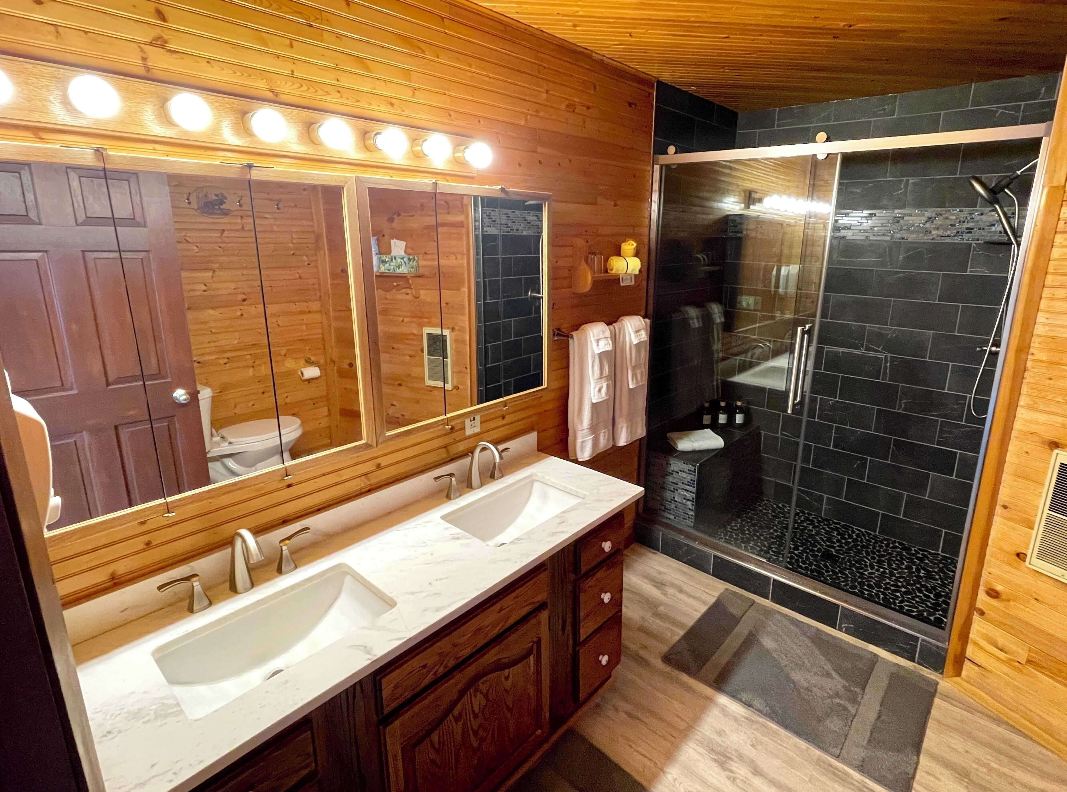 Un baño con paredes de madera, un lavabo doble, un espejo grande con iluminación y una ducha con azulejos negros, todo bajo un techo de madera.