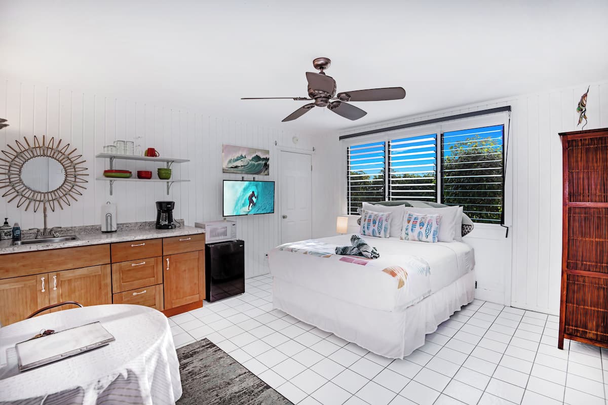 A bedroom with a bed, a television, a ceiling fan, a window with blinds, and a wooden cabinet.
