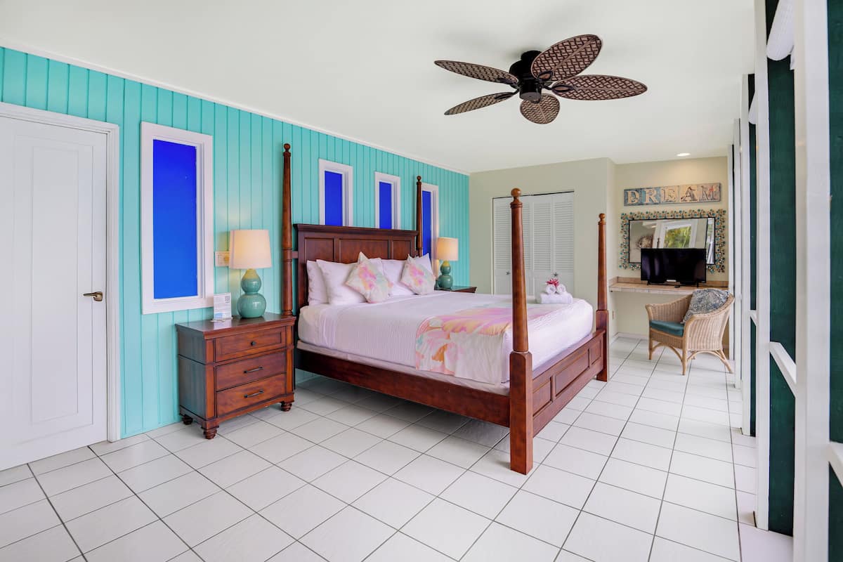 A four-poster bed with a wooden frame, a nightstand with a lamp, and a chair in a room with teal walls and white tiled flooring.