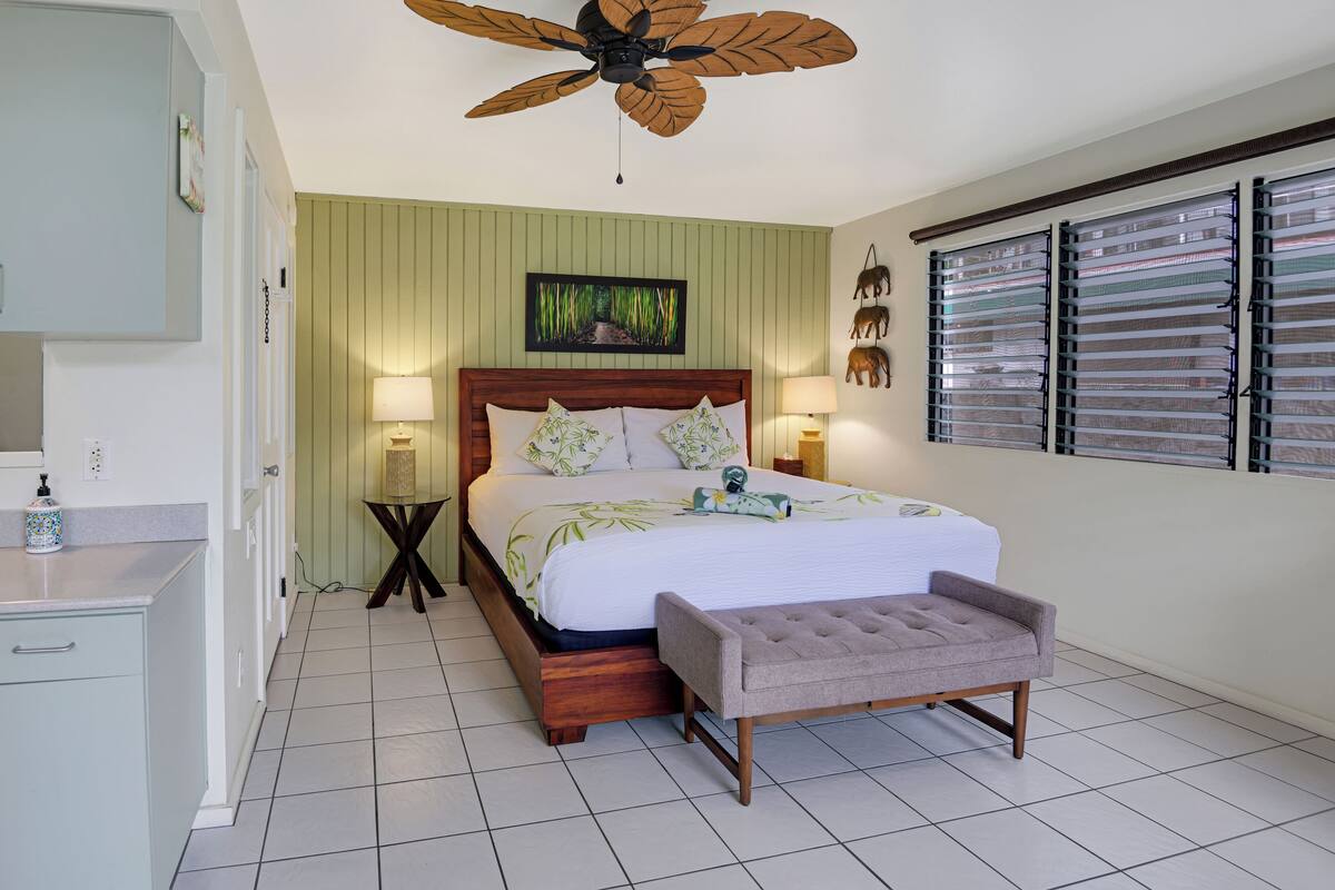 A bedroom with a bed, bedside lamps, a ceiling fan, a window with blinds, and a small table with a lamp.