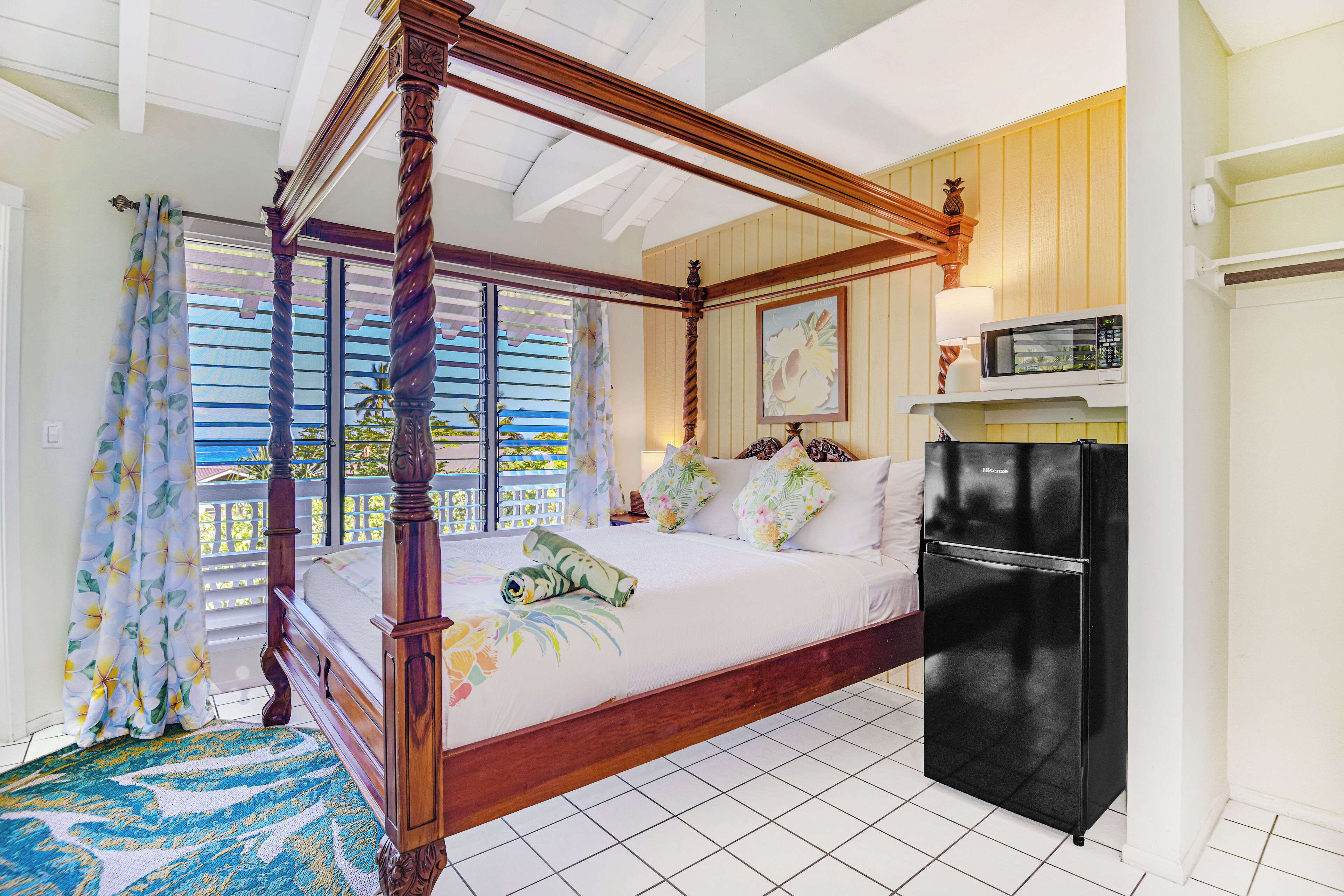 A four-poster bed with a wooden frame, a black refrigerator, and a view of the sea through the window.