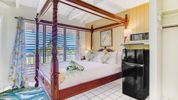 A four-poster bed with a wooden frame, a black refrigerator, and a view of the sea through the window.