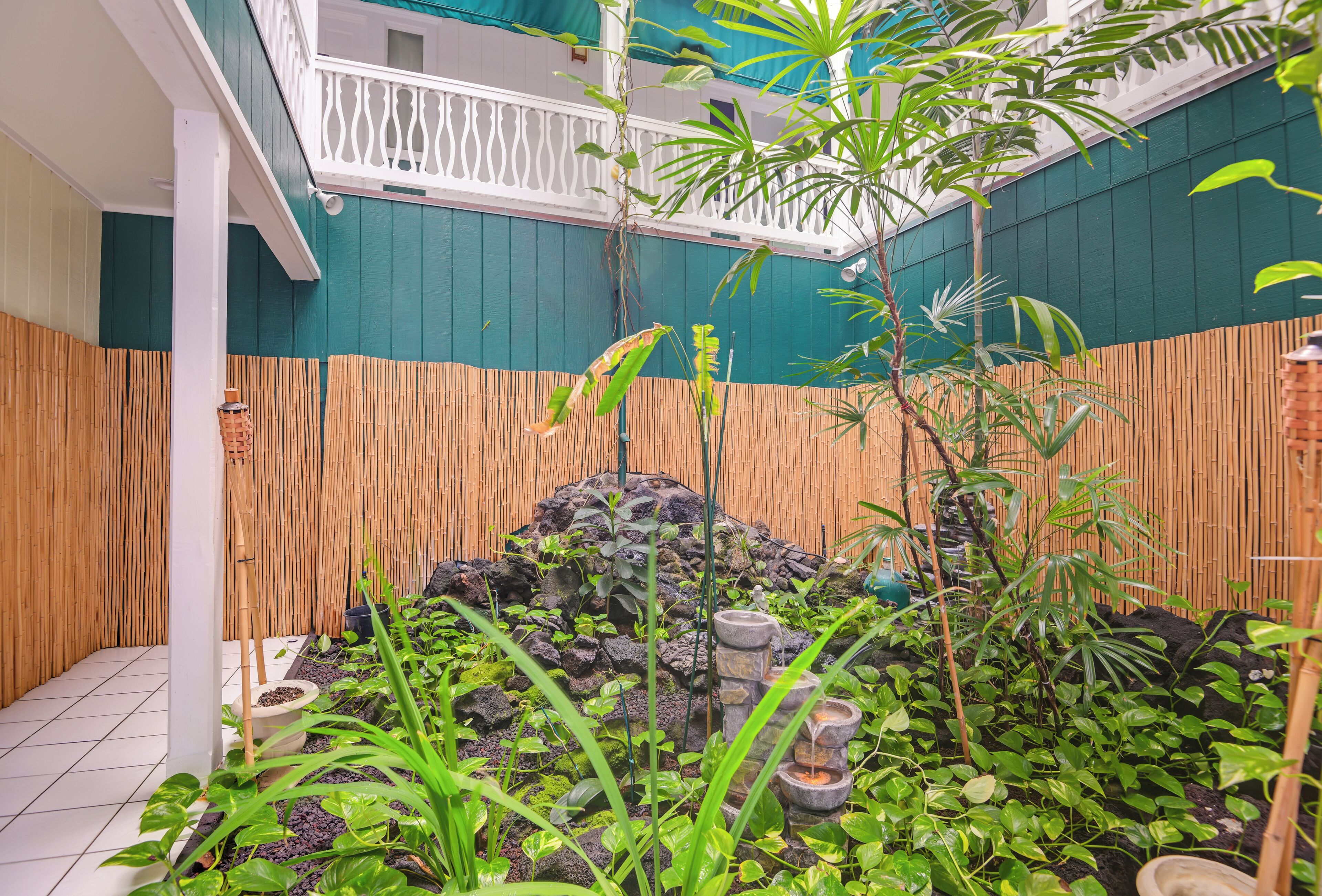 A small, enclosed garden area with a variety of plants, a small rock formation, and a bamboo fence.