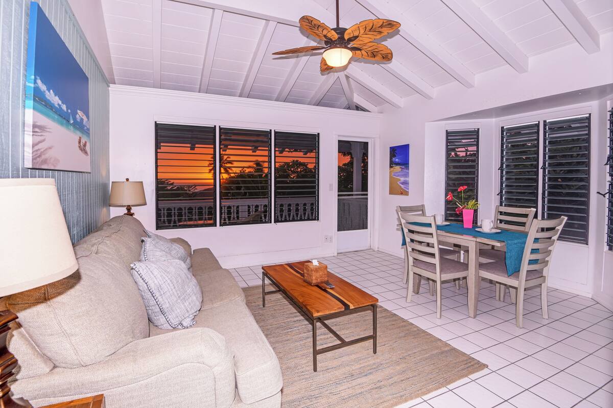 A living room with a sofa, coffee table, dining table with chairs, a ceiling fan, and large windows with shutters.