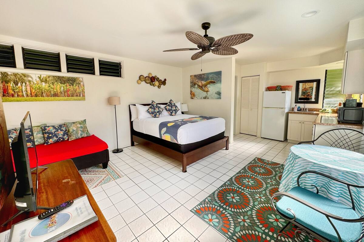 A compact hotel room with a bed, a desk, a chair, a ceiling fan, and a kitchenette.