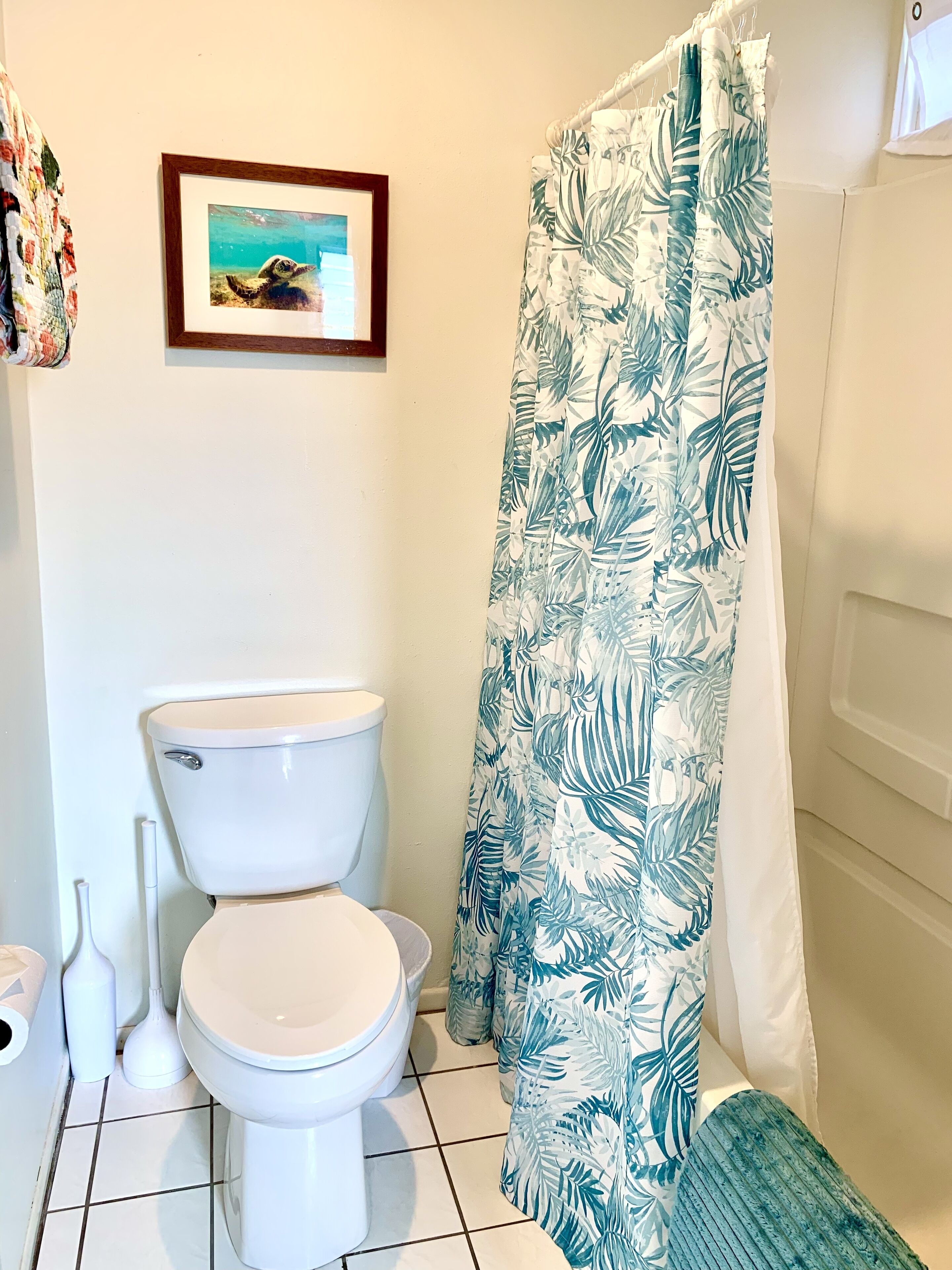 Une salle de bain avec des toilettes, un rideau de douche orné d’un motif de feuilles et un tableau encadré accroché au mur.