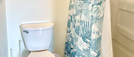 Une salle de bain avec des toilettes, un rideau de douche orné d’un motif de feuilles et un tableau encadré accroché au mur.