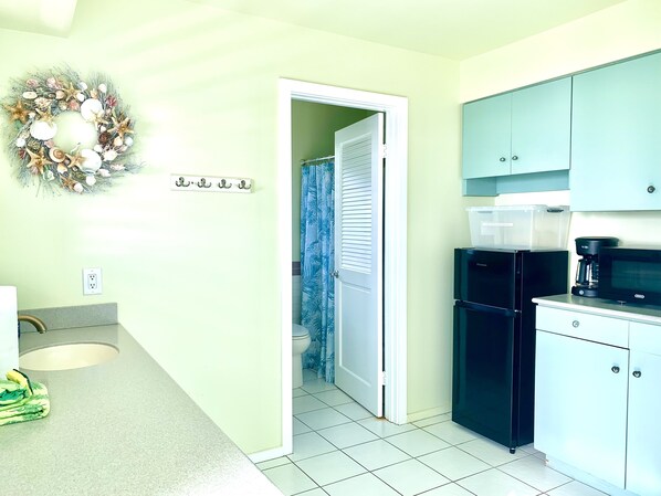 A kitchen with a refrigerator, microwave, and a coffee maker. There is a door leading to a bathroom with a shower curtain.