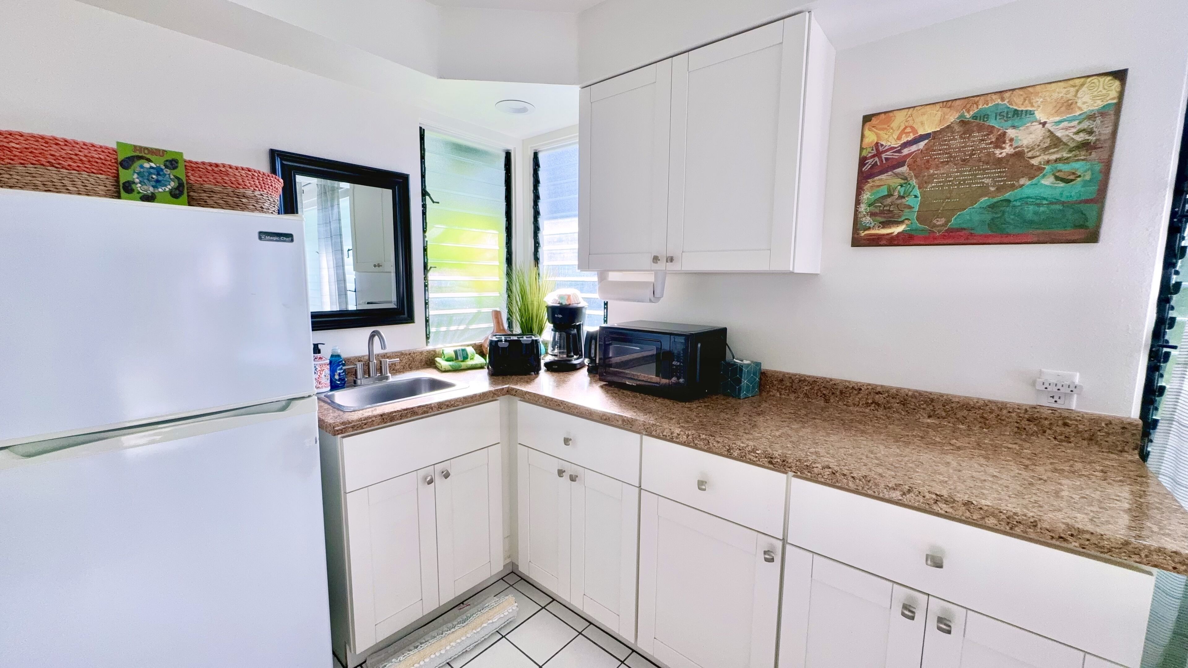 A compact kitchen with white cabinets, a granite countertop, a refrigerator, a sink, a coffee maker, a toaster, and a microwave.