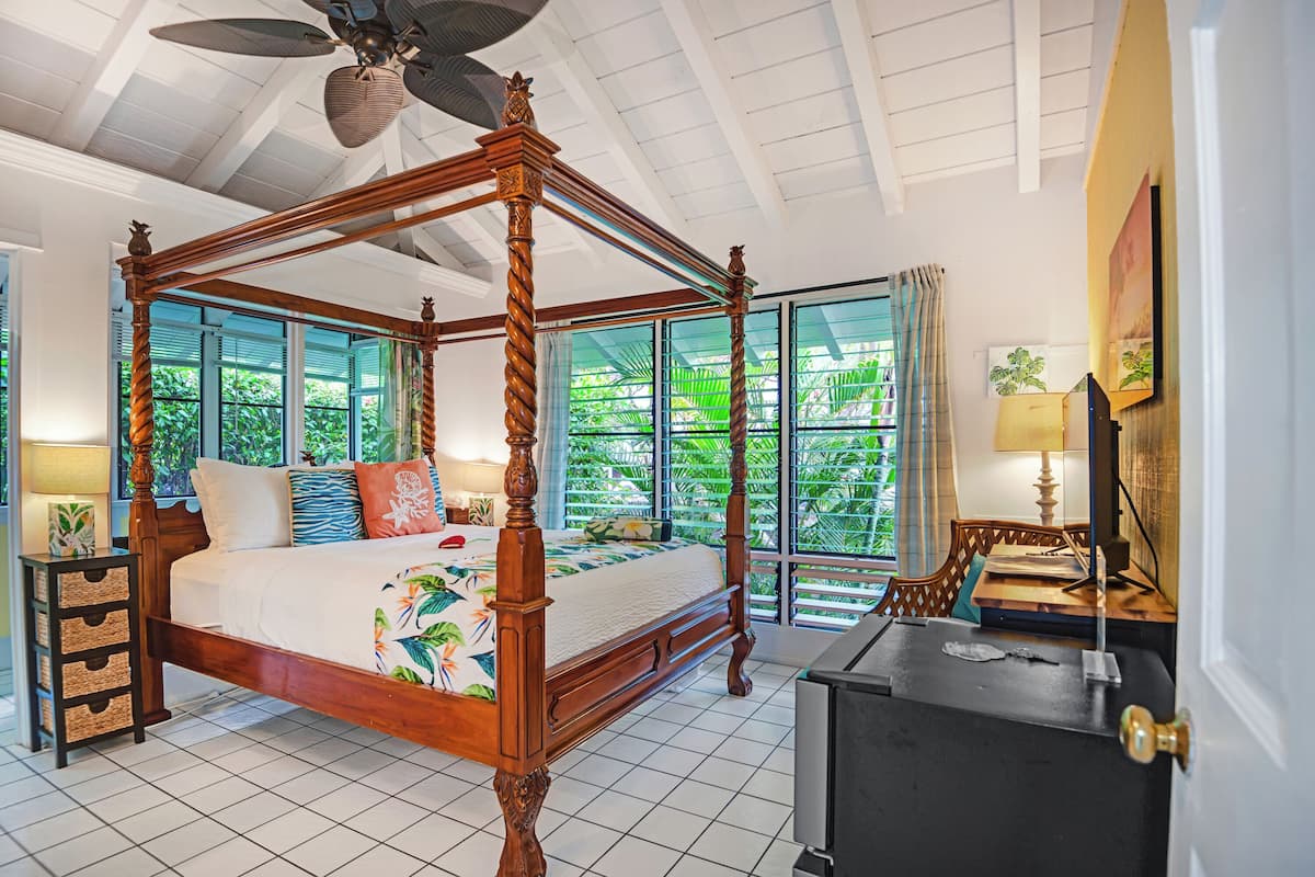 A four-poster bed with a tropical-themed bedspread, a wooden nightstand with drawers, a desk with a lamp, and a large window with a view of greenery.