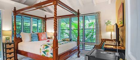 Een hemelbed met een tropisch beddegoed, een nachtkastje van hout met lades, een bureau met een lamp en een groot raam met uitzicht op groen.