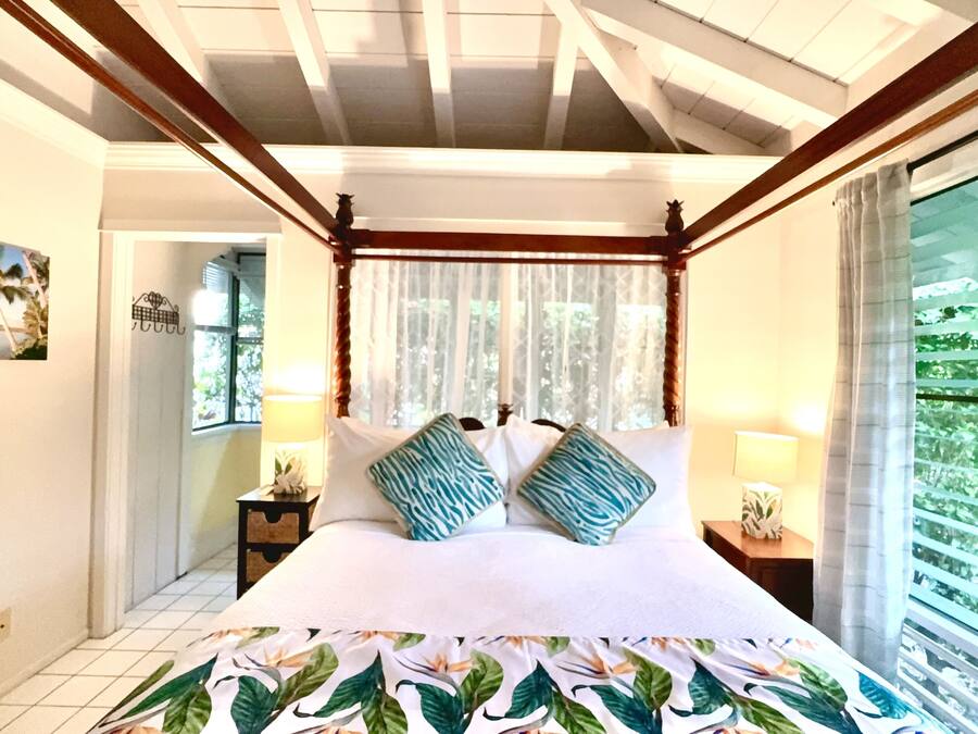 A bedroom with a bed, a nightstand, a lamp, a window with curtains, and a view of greenery outside.