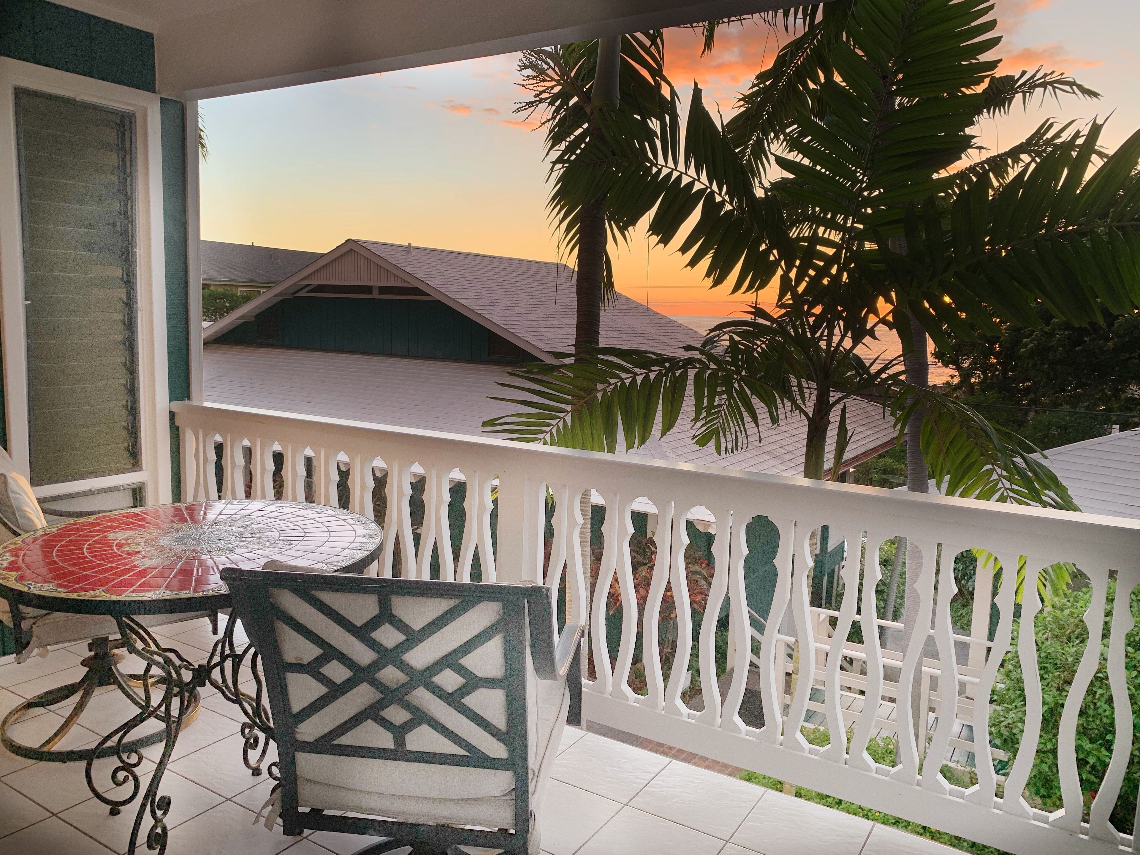 A balcony with a table and chair, overlooking a residential area and a sunset.