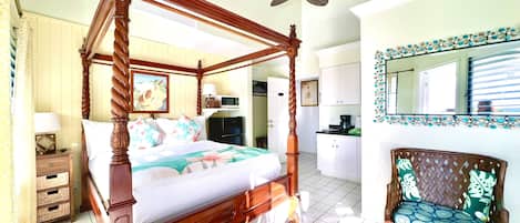 A four-poster bed with a floral bedspread, a wooden nightstand with a lamp, a wicker chair with a cushion, and a kitchen area with a microwave and cabinets.