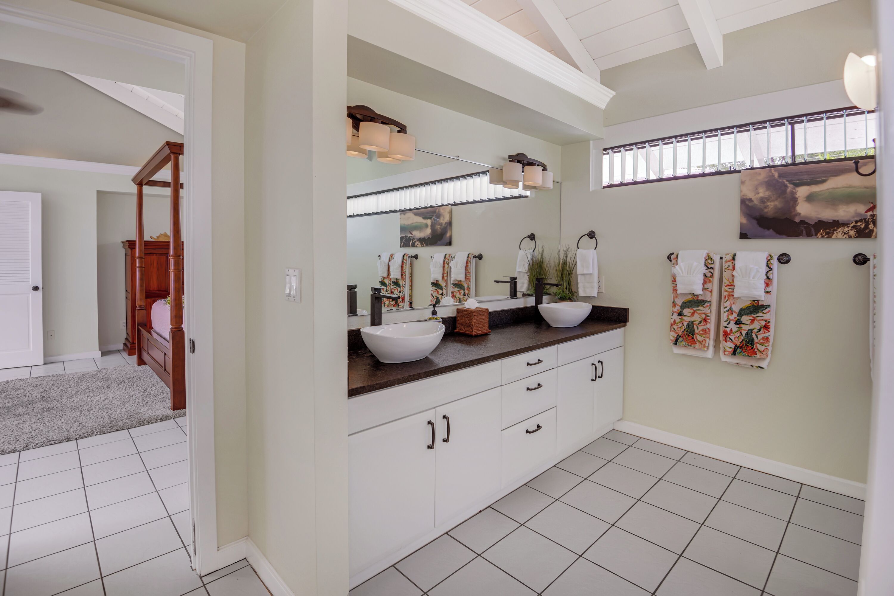 A bathroom with a double sink, white cabinets, and a large mirror. There are towels hanging on the wall and a window with blinds.