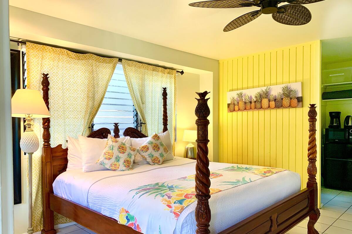 A four-poster bed with a canopy, a bedside lamp, and a pineapple-themed pillow.