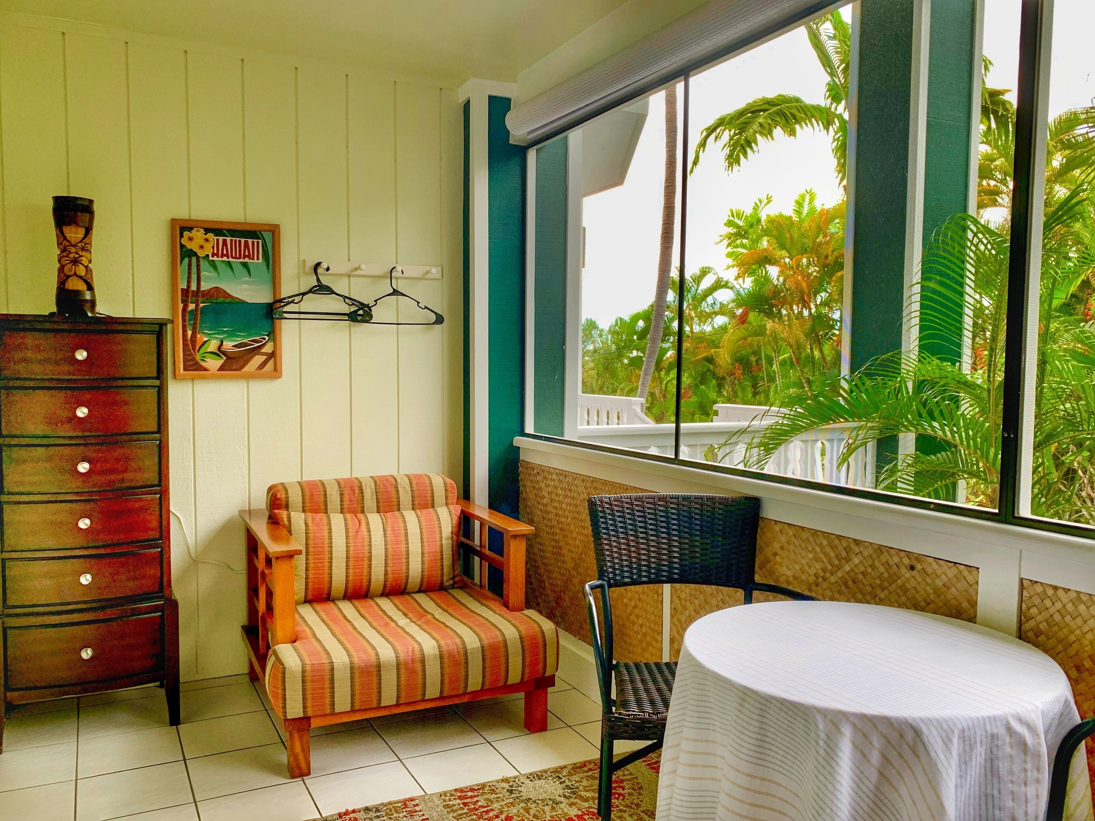 A room with a wooden dresser, a striped sofa, a wicker chair, and a small table. A window offers a view of palm trees and a white fence.