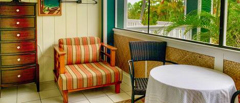 A room with a wooden dresser, a striped sofa, a wicker chair, and a small table. A window offers a view of palm trees and a white fence.