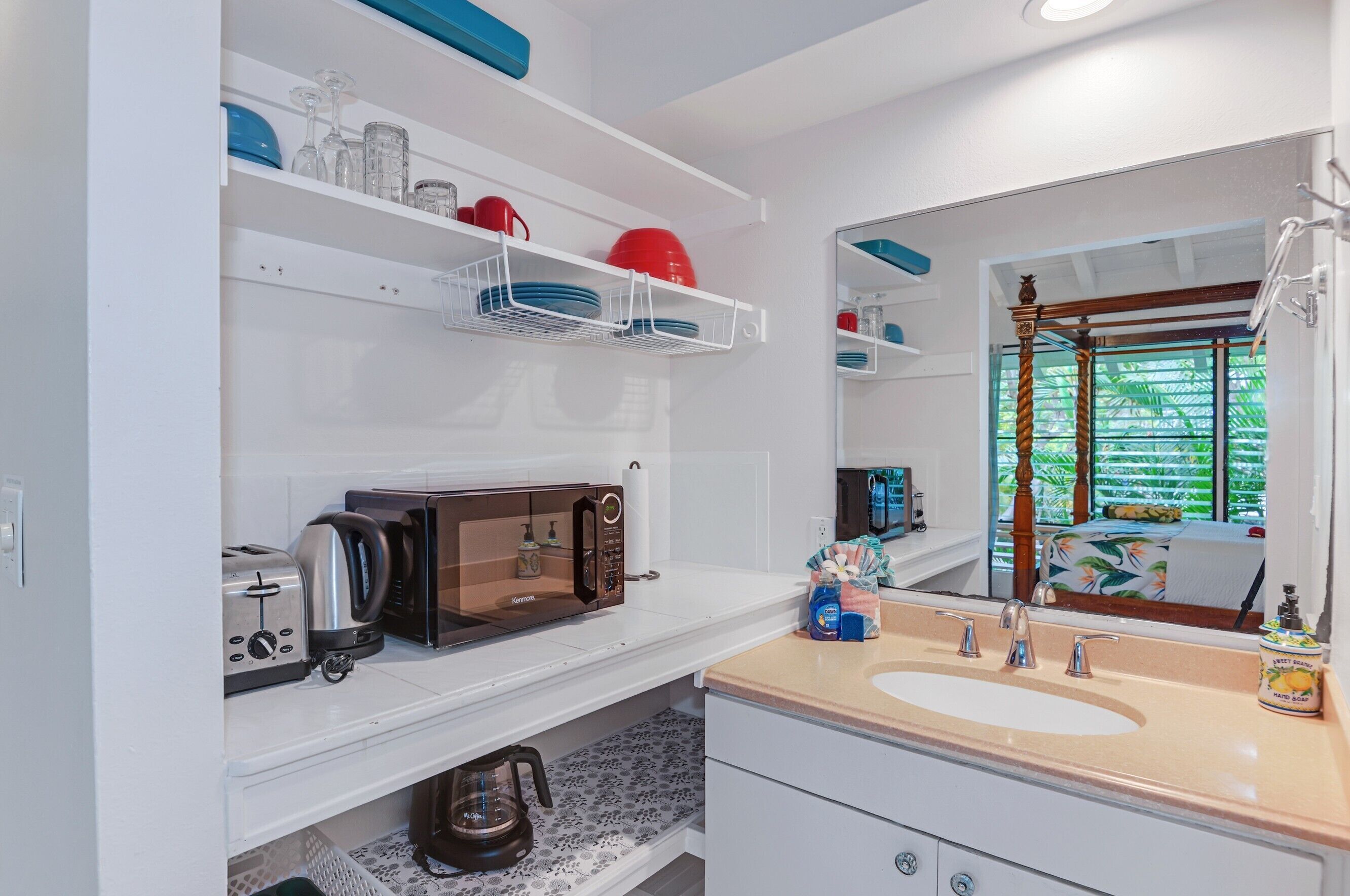 A modern bathroom with a large mirror, a sink, a microwave, and shelves with dishes and toiletries.
