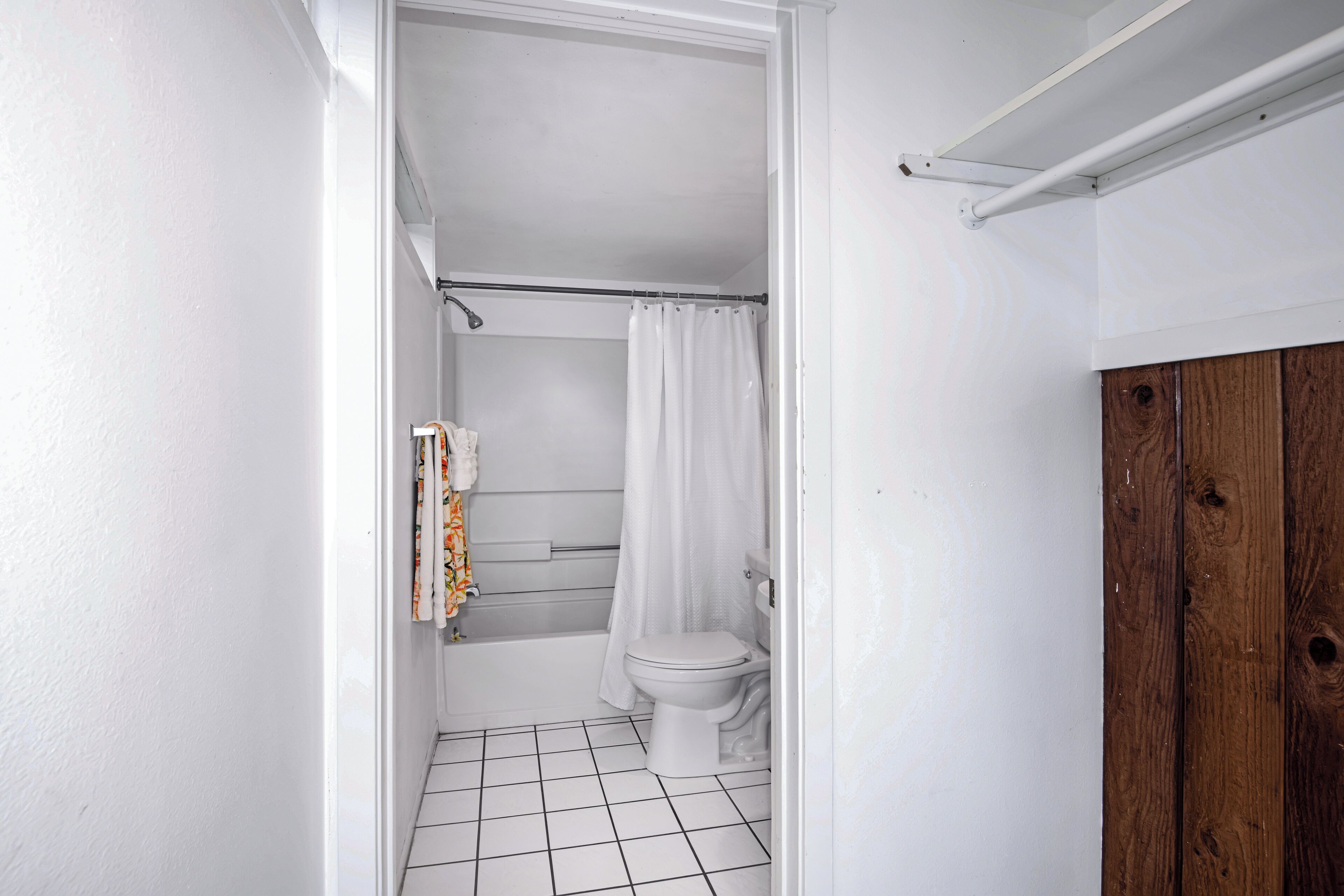 Une petite salle de bain aux murs blancs, avec des toilettes blanches, une douche dotée d’un rideau blanc et un meuble en bois.