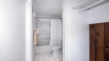 Une petite salle de bain aux murs blancs, avec des toilettes blanches, une douche dotée d’un rideau blanc et un meuble en bois.