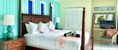 A four-poster bed with a wooden headboard, two nightstands with lamps, a television on a stand, and a wicker chair with a cushion.