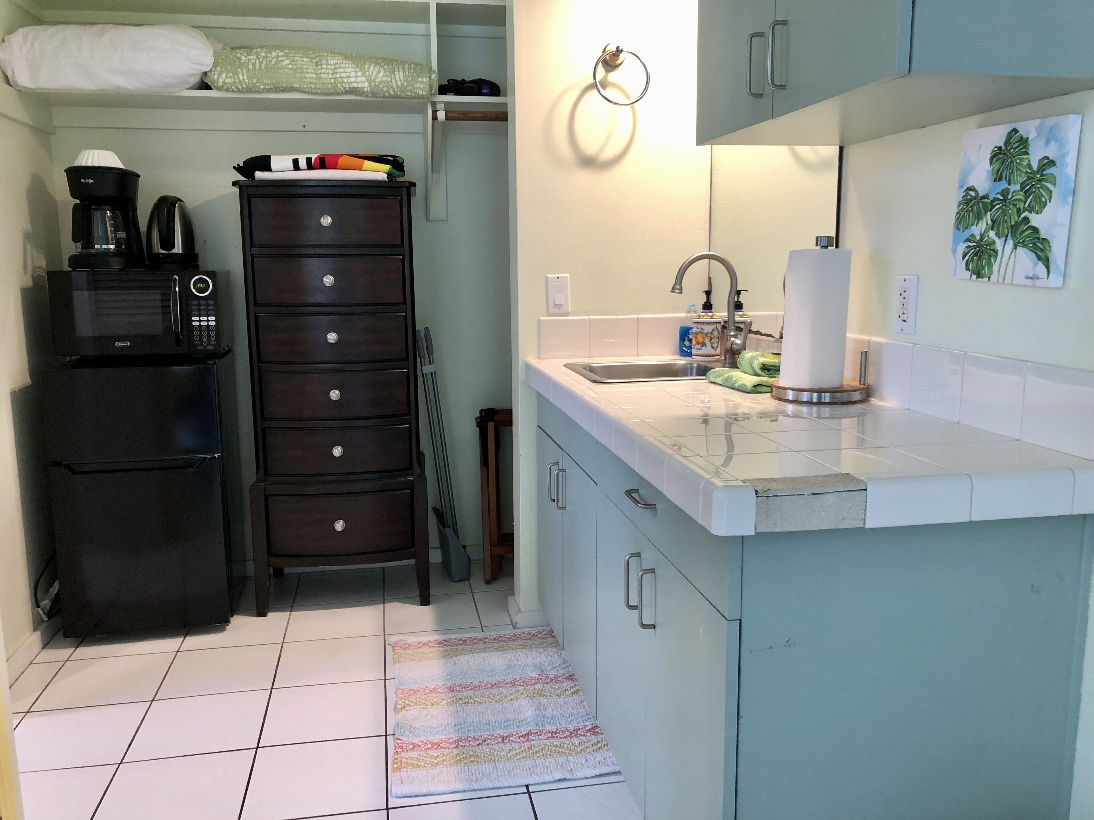 A compact kitchen with a refrigerator, microwave, and coffee maker. There is a sink with a faucet, a towel dispenser, and a small cabinet. A colorful rug is on the floor.