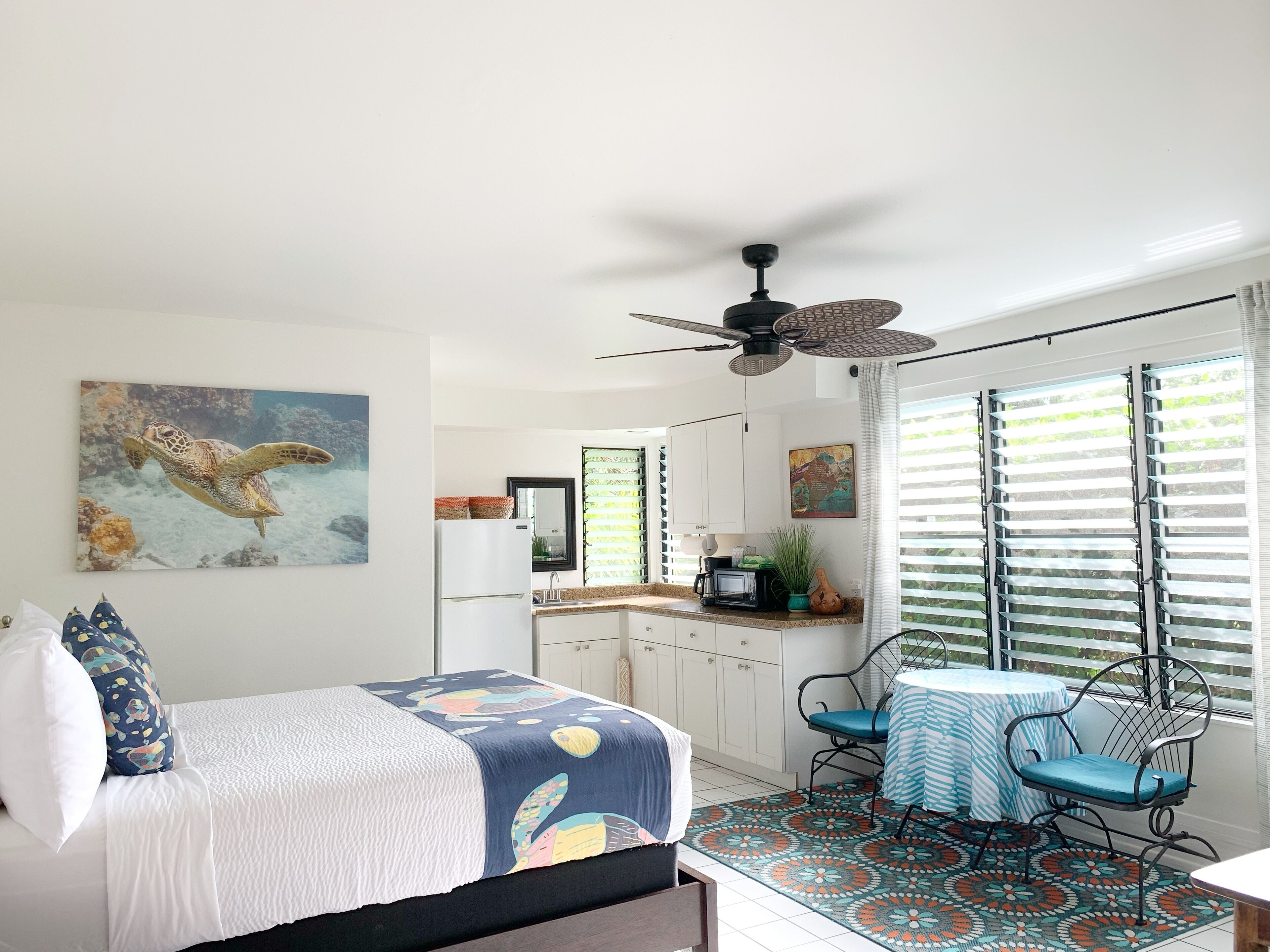 Une chambre à coucher comprenant un lit, un ventilateur de plafond, un coin cuisine avec des placards et un réfrigérateur, ainsi qu’un coin repas avec une table et des chaises.
