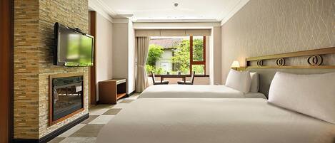 A hotel room with a large bed, a flat-screen TV mounted on a stone wall, a fireplace, and a view of greenery outside.