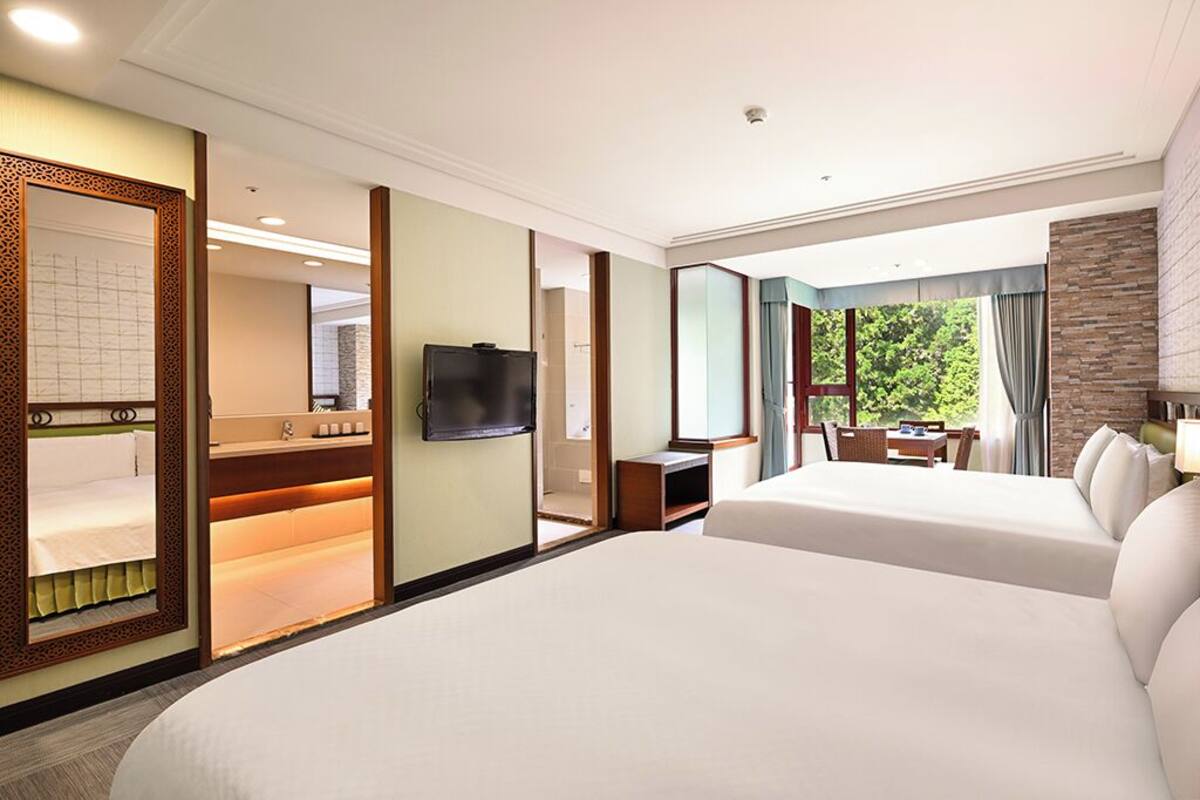 A hotel room with two beds, a TV, a desk, and a view of greenery.