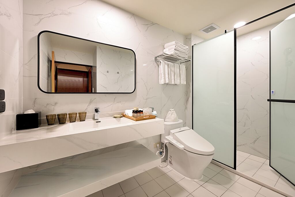 A modern bathroom with a marble wall, a large mirror, a toilet, a sink with toiletries, and a glass shower enclosure.