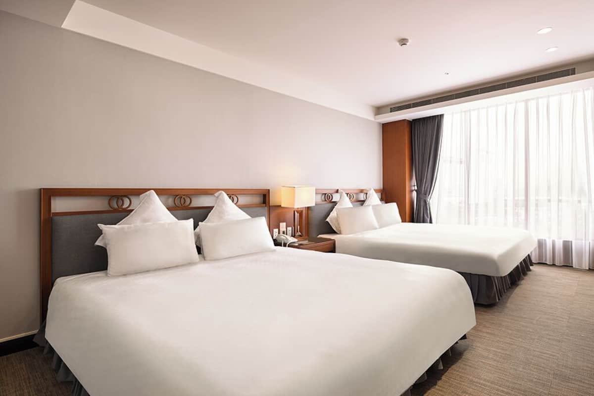 A hotel room with two beds, a nightstand with a lamp, a large window with curtains, and a ceiling vent.