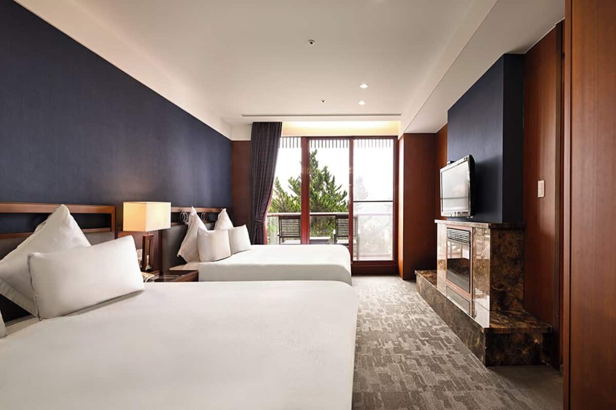 A hotel room with two beds, a TV, a fireplace, and a balcony with a view of trees.
