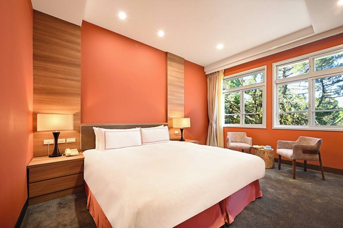 A hotel room with a large bed, two bedside tables, a nightstand, two chairs, and a window with a view of trees.