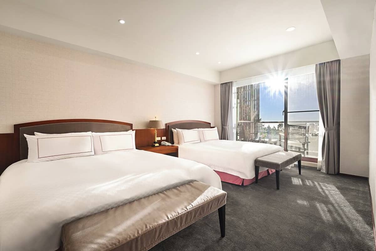 A hotel room with two beds, a bench, a nightstand, and a large window with a city view.