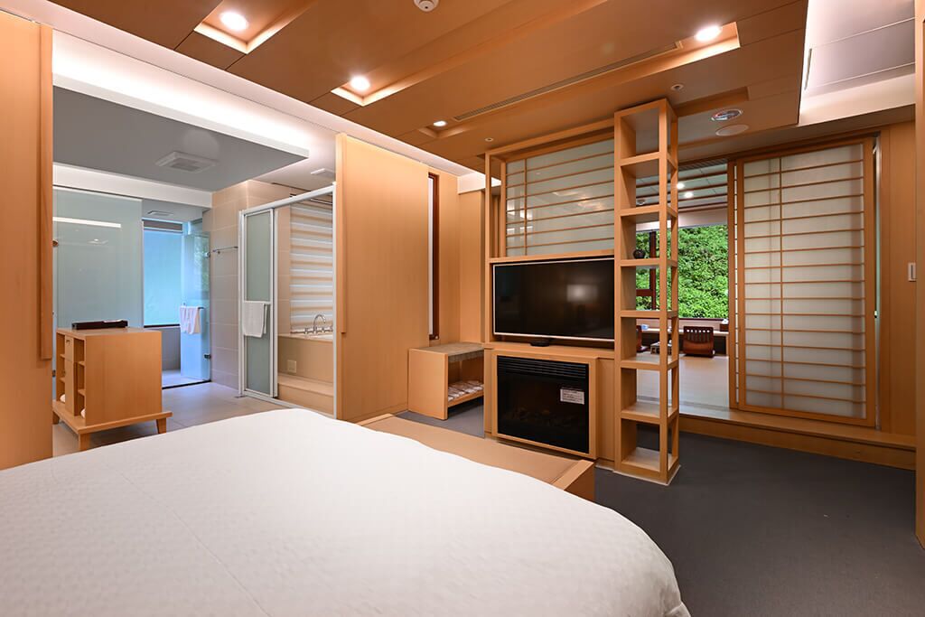 A modern hotel room with a large bed, a TV, sliding doors, and a view of greenery.
