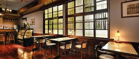 Un restaurante con suelo de madera, varias mesas y sillas, ventanas amplias con barandillas y candelabros colgantes.