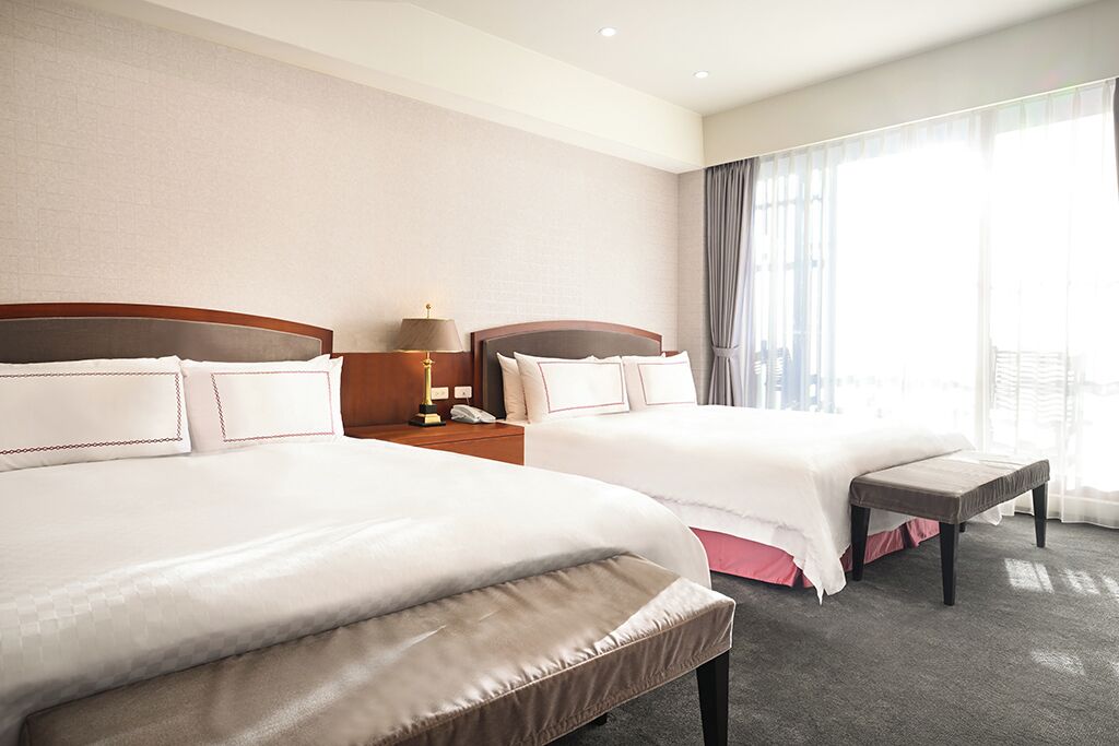A hotel room with two beds, a nightstand with a lamp, and a large window with curtains.