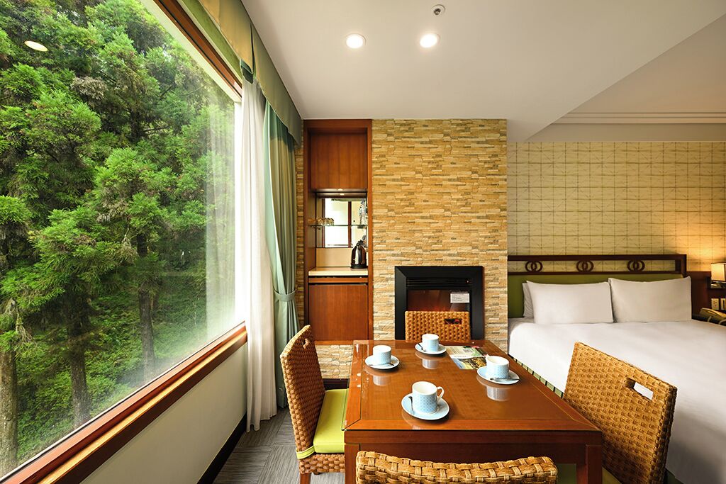 A hotel room with a large window offering a forest view, a wooden dining table with chairs, a bed with white linens, and a stone fireplace.