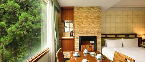 A hotel room with a large window offering a forest view, a wooden dining table with chairs, a bed with white linens, and a stone fireplace.