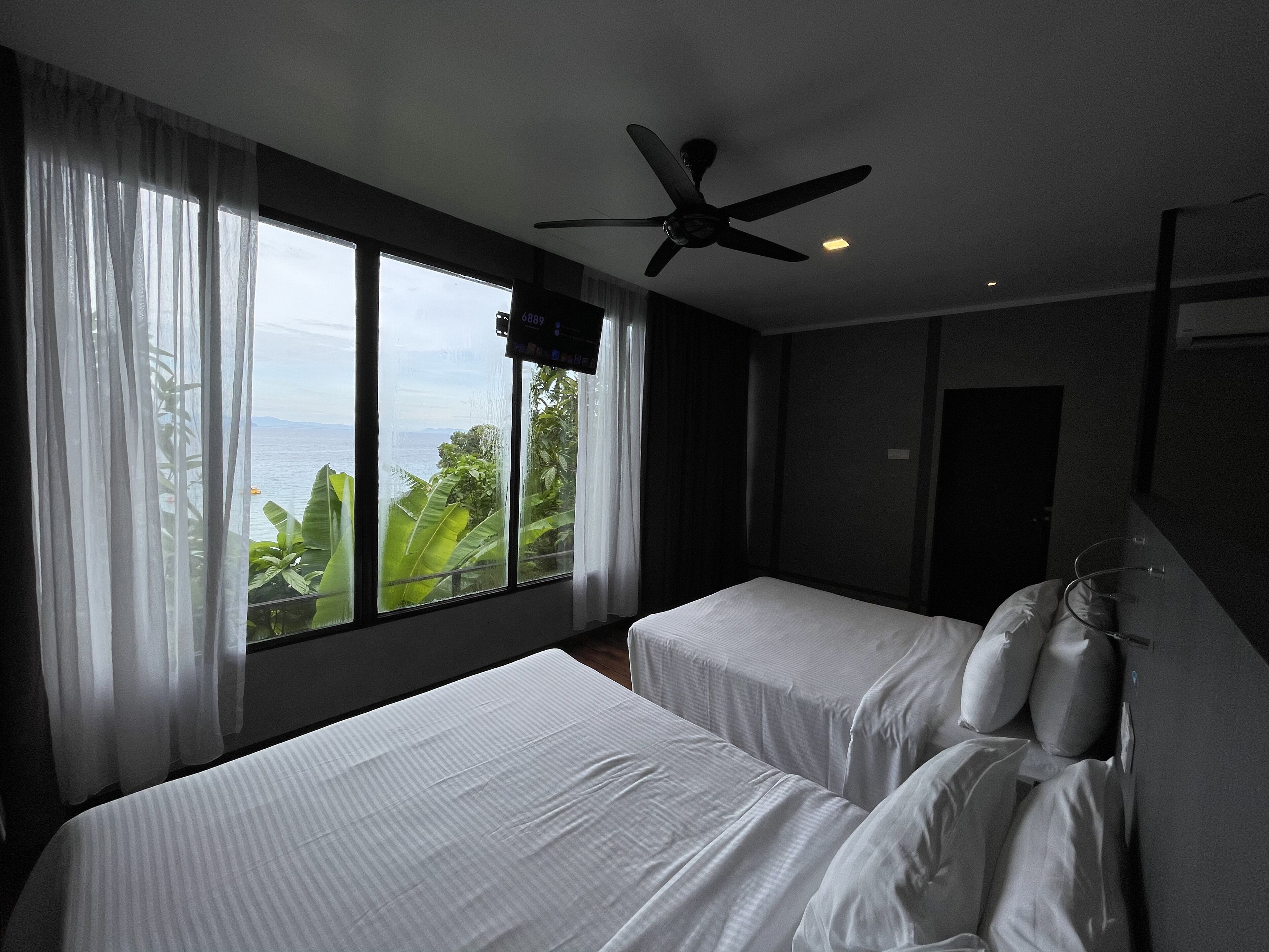 Habitación de hotel con dos camas, ventilador de techo, televisión y un amplio ventanal con cortinas.
