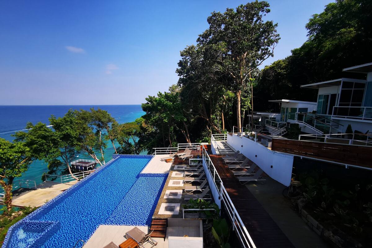 A rooftop infinity pool with a view of the ocean, surrounded by lush greenery and modern outdoor seating.